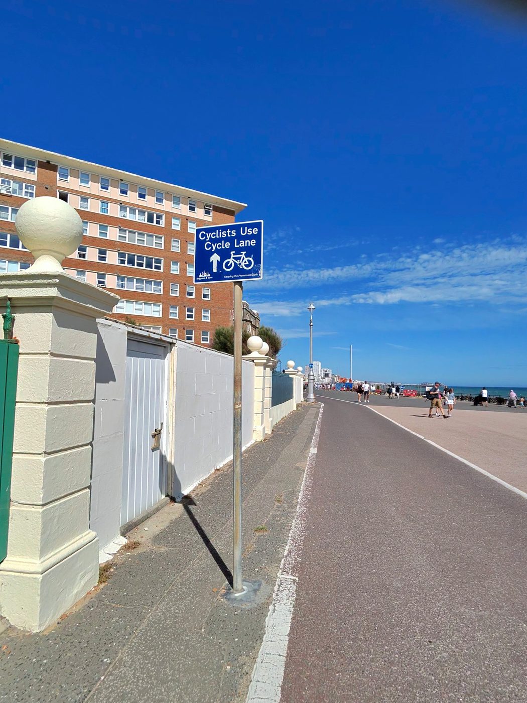 A real Brighton and Hove council sign on the seafront promenade reading Cyclists Use Cycle Lane, with an arrow and bicycle symbol above the strapline Keeping the Promenade Safe.